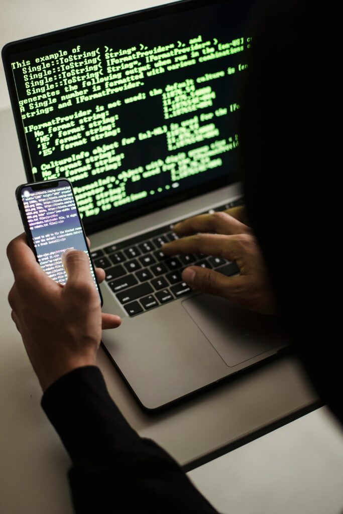 About Us A hooded figure engaged in hacking using a laptop and smartphone in low light.